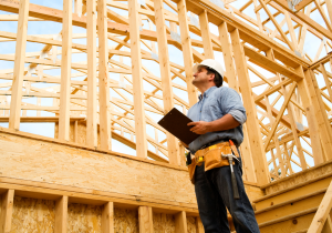 man with clipboard, toolbelt, and hardhat looking up at wood frame construction