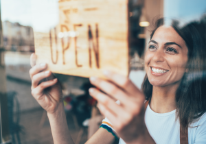 smiling woman turning sign in window around so that "Open" faces outside