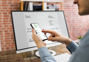 man looking at purchase receipt on smartphone with desktop computer with invoice in background